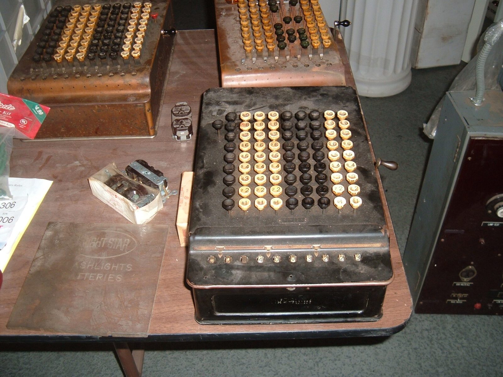Three vintage mechanical adding machines with rows of round number keys on a table at a flea market, beside boxes of old office supplies.