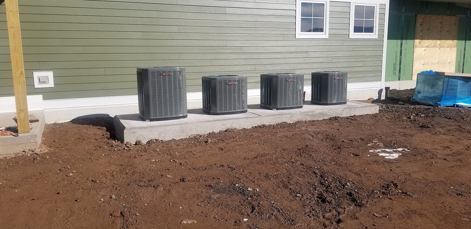 Four new outdoor air-conditioning condenser units set in a row on a concrete pad beside a building with green siding, with fresh soil in the foreground.