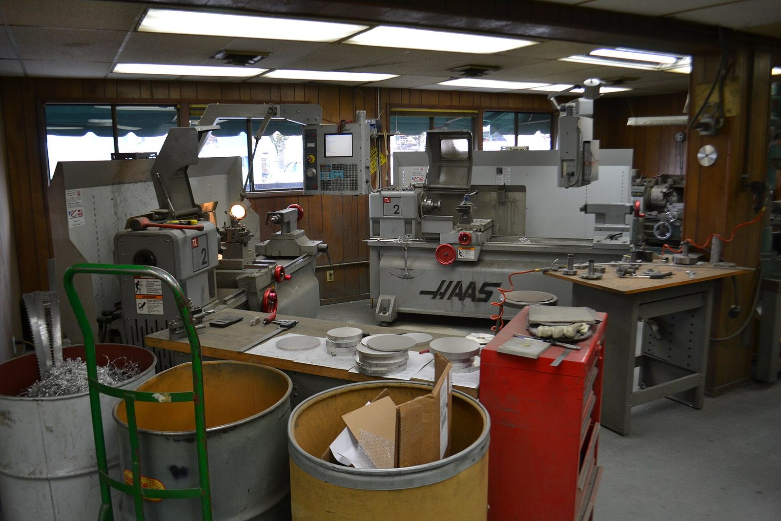 Interior of a small machine shop with gray Haas lathes along wood-paneled walls, a red tool chest, a green hand truck and barrels of metal scrap under fluorescent lights.