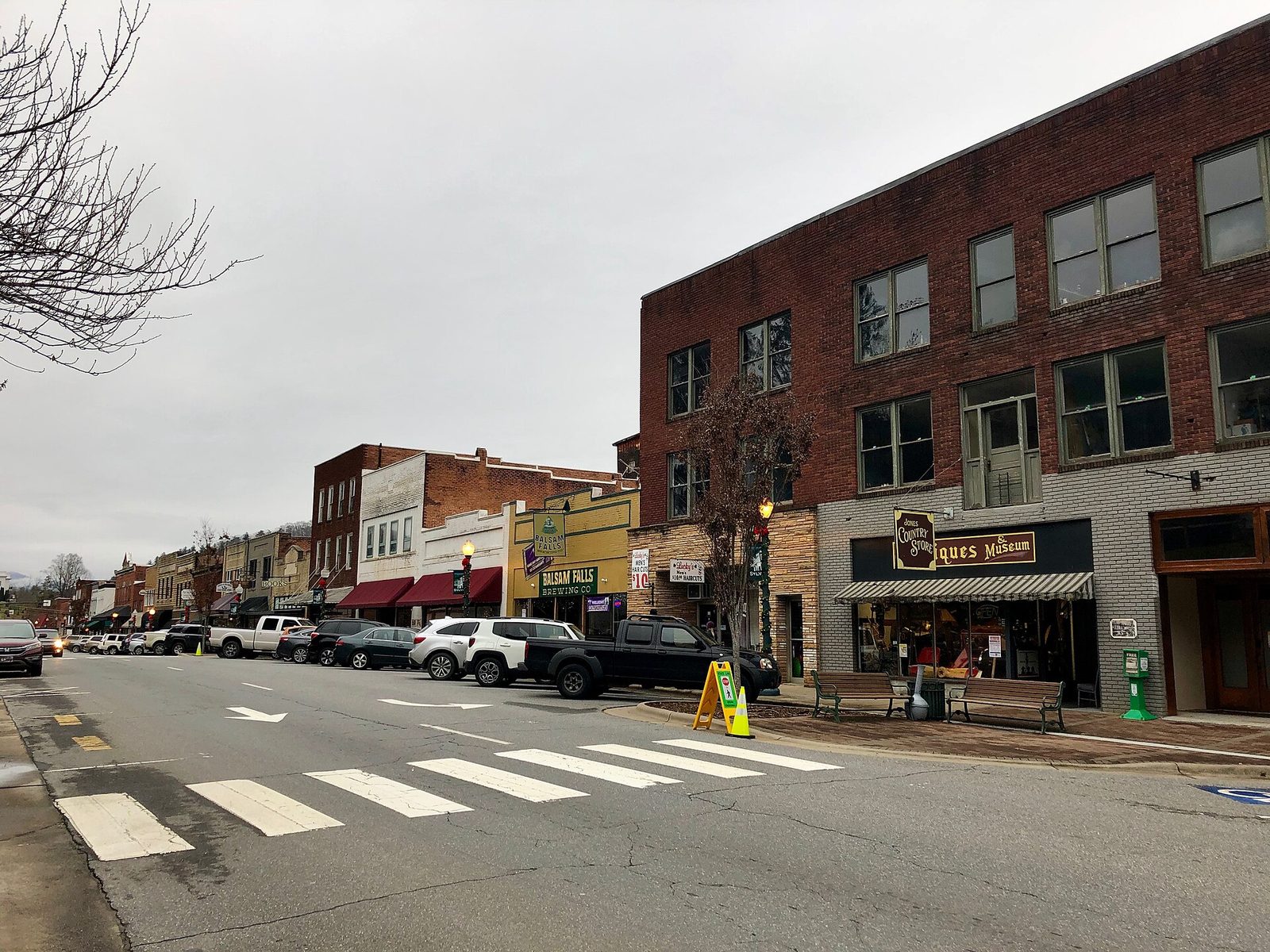 Main Street in Sylva, North Carolina, with a three-story brick building on the corner, a crosswalk and a row of small shops and parked trucks under an overcast sky.