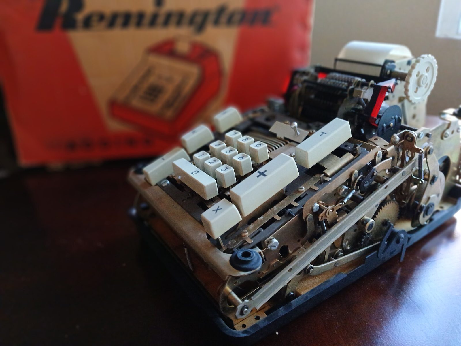 Close view of a vintage Remington adding machine with its cover removed, showing the number keys and gears, in front of its red and white cardboard box.