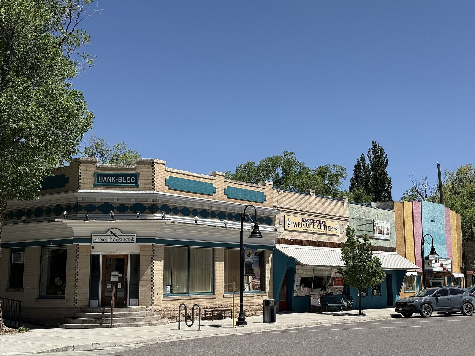 A low corner bank building with a sign reading Bank Bldg beside a welcome center and other one-story storefronts on a sunny small-town street in Saguache, Colorado.