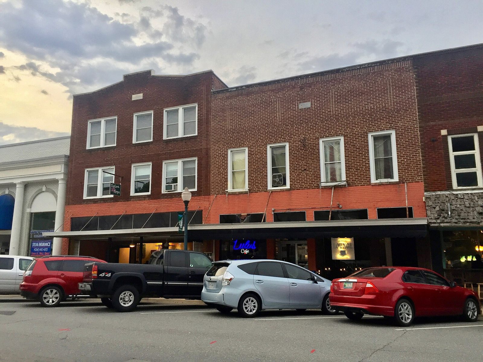 A row of 1920s two- and three-story brick commercial buildings on Main Street in Sylva, North Carolina, with storefront windows, awnings and cars parked along the curb at dusk.