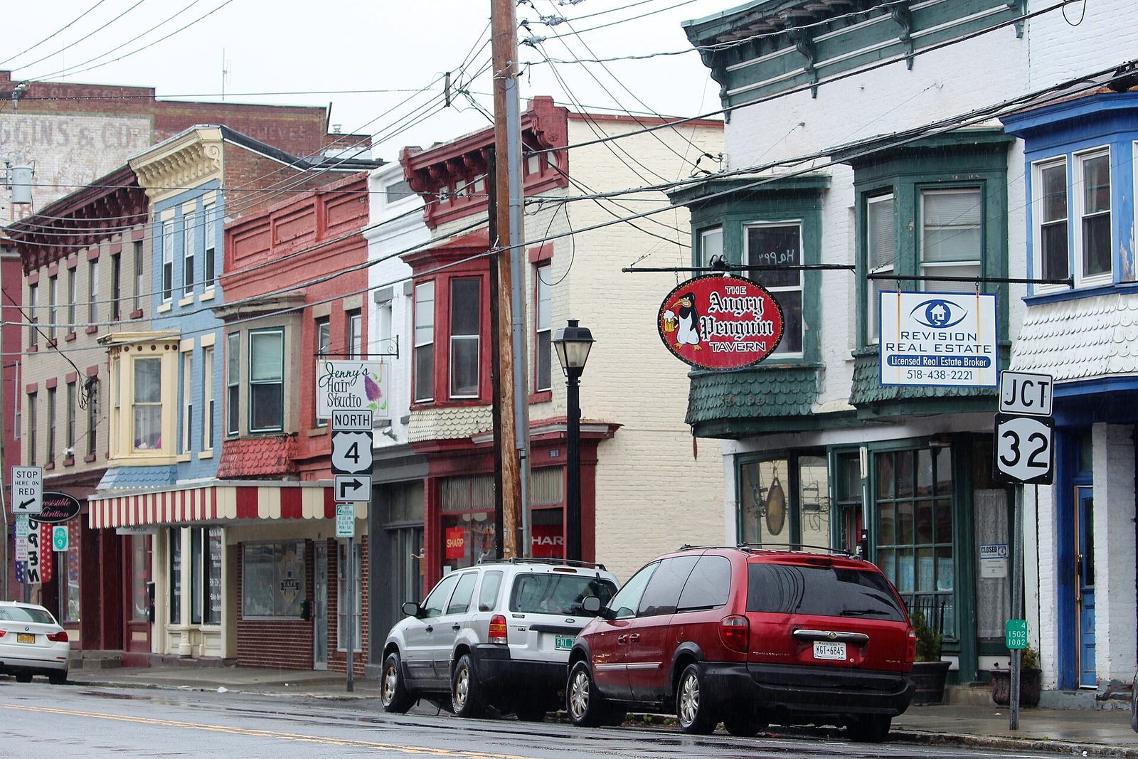 Broad Street in Waterford, New York: two- and three-story painted storefront buildings with bay windows, hanging shop signs and a route marker at the corner.